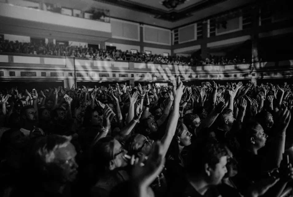 Large crowd of people with raised hands at an indoor concert venue with balcony seating.