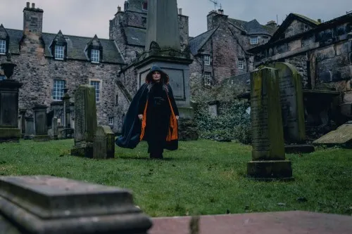 Person in dark clothing walking among historic gravestones with stone buildings in background.