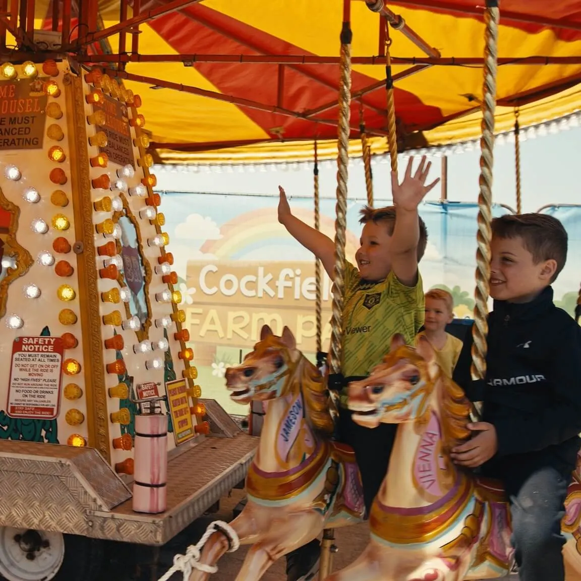 Children riding a traditional carousel with painted horses under a striped awning at a farm park.