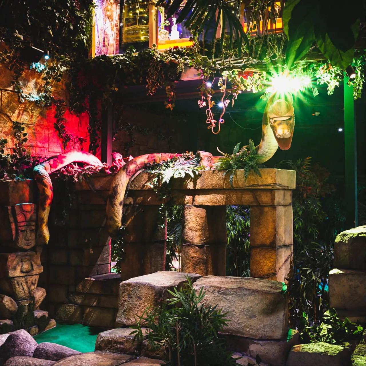 Colourful indoor mini golf course with jungle-themed stone structures, tropical plants, and neon lighting in red, green and purple.