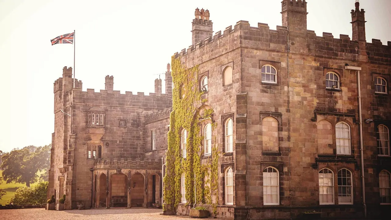 Historic red sandstone castle with crenellated walls, ivy-covered tower, multiple chimneys, arched windows and a Union Jack flag flying from the battlements.