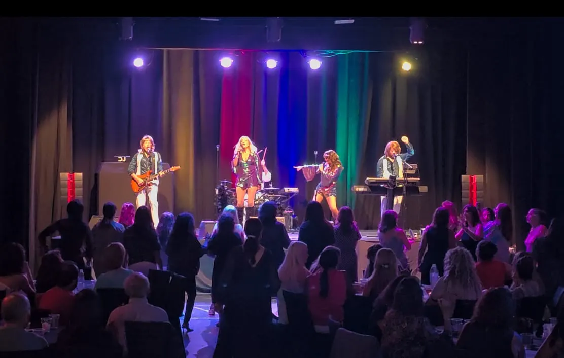 Four musicians performing on stage with colourful lighting, watched by a seated audience in an indoor venue.
