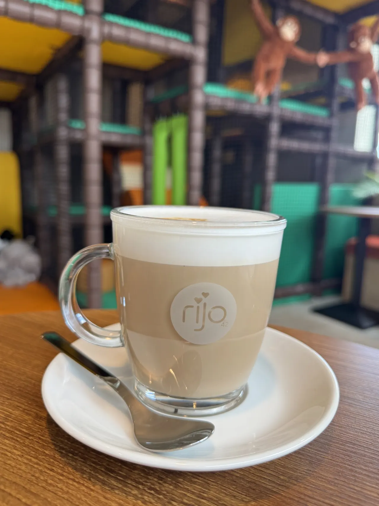Cappuccino with latte art in a glass mug on a saucer at an indoor play centre with climbing structures visible in the background.