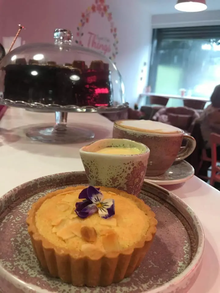 A yellow tart with edible flowers sits on a plate beside two cups of tea, with a cake under a dome visible behind.