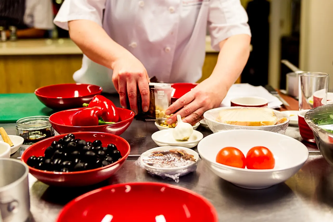 A chef prepares ingredients at a cooking class, arranging red peppers, tomatoes, olives and other fresh produce in bowls.