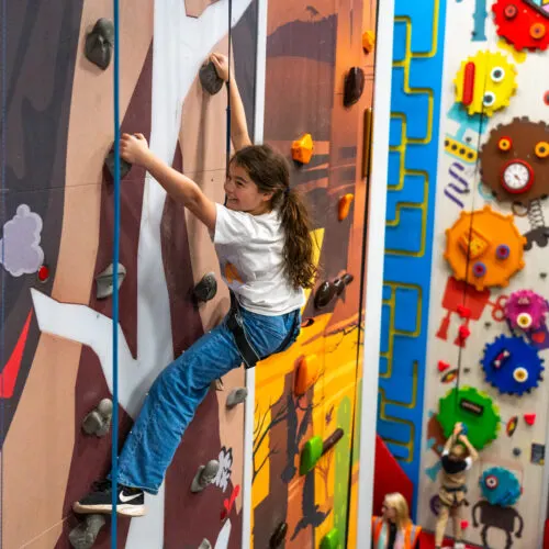 Child climbing an indoor climbing wall with coloured handholds and painted designs.