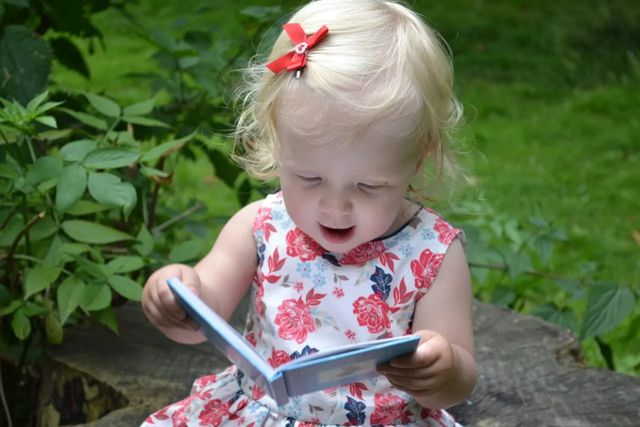 Toddler with blonde hair and red bow looking at a blue handheld device in a garden surrounded by green foliage.