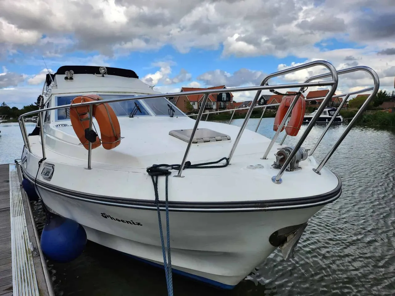 White motorboat with orange life rings moored at a jetty, red-brick buildings visible across the water under a cloudy sky.