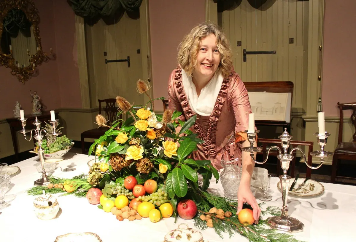 Woman posing beside an elaborate formal dining table arrangement of yellow flowers, fresh fruits and greenery with candelabras.