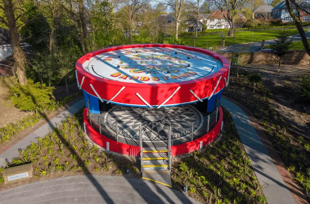 Aerial view of a large red and blue rotating carousel with decorative panels, set in a tree-lined park with residential houses visible beyond.