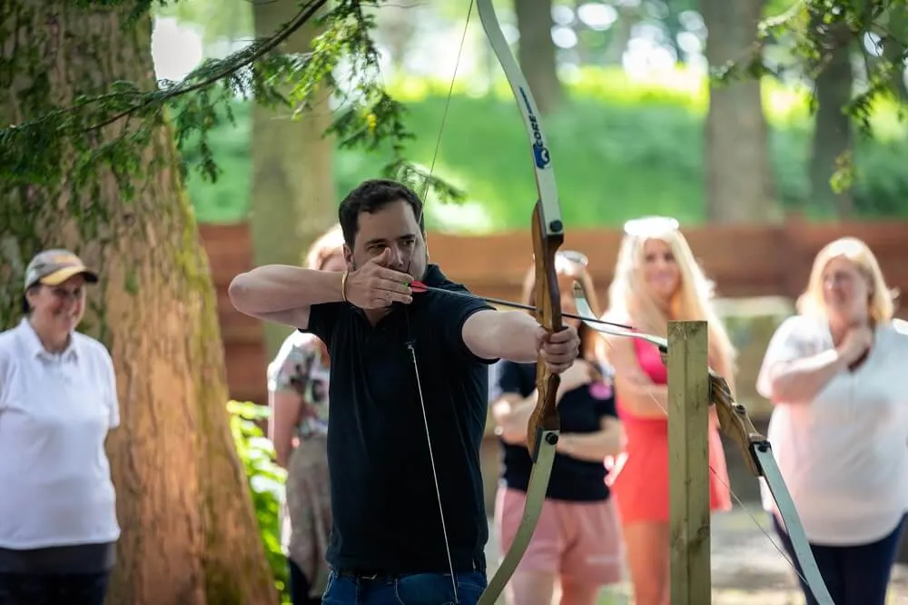 Man in black shirt drawing a bow and arrow whilst spectators watch in a woodland setting.