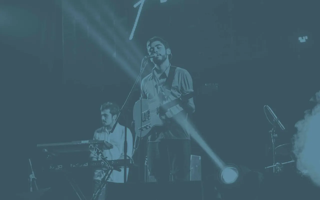 A band performs on stage in a dimly lit venue with blue stage lighting, featuring musicians playing guitar and keyboards.