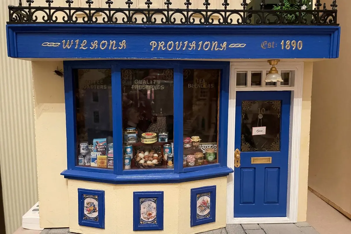 A detailed miniature model of a blue and cream Victorian shopfront displaying vintage provisions and groceries in its windows.