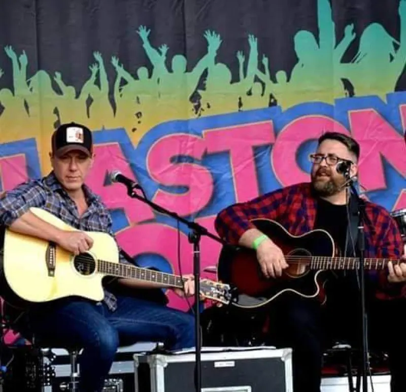 Two men perform with acoustic guitars on an outdoor stage with colourful graffiti-style backdrop.