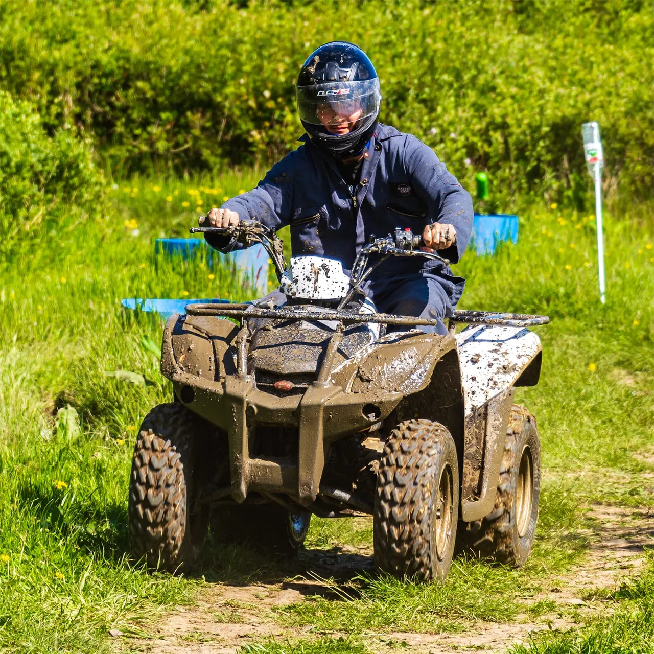 Person in helmet riding a mud-covered quad bike through a grassy outdoor course with blue markers.