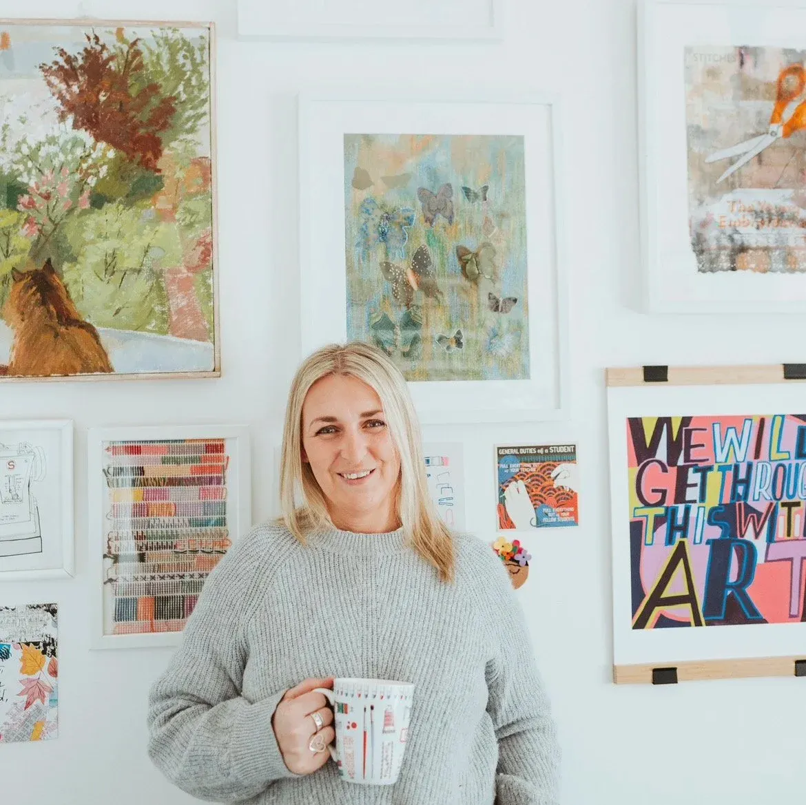 A woman in a grey sweater holding a mug stands smiling in front of a white wall displaying various framed artworks and illustrations.