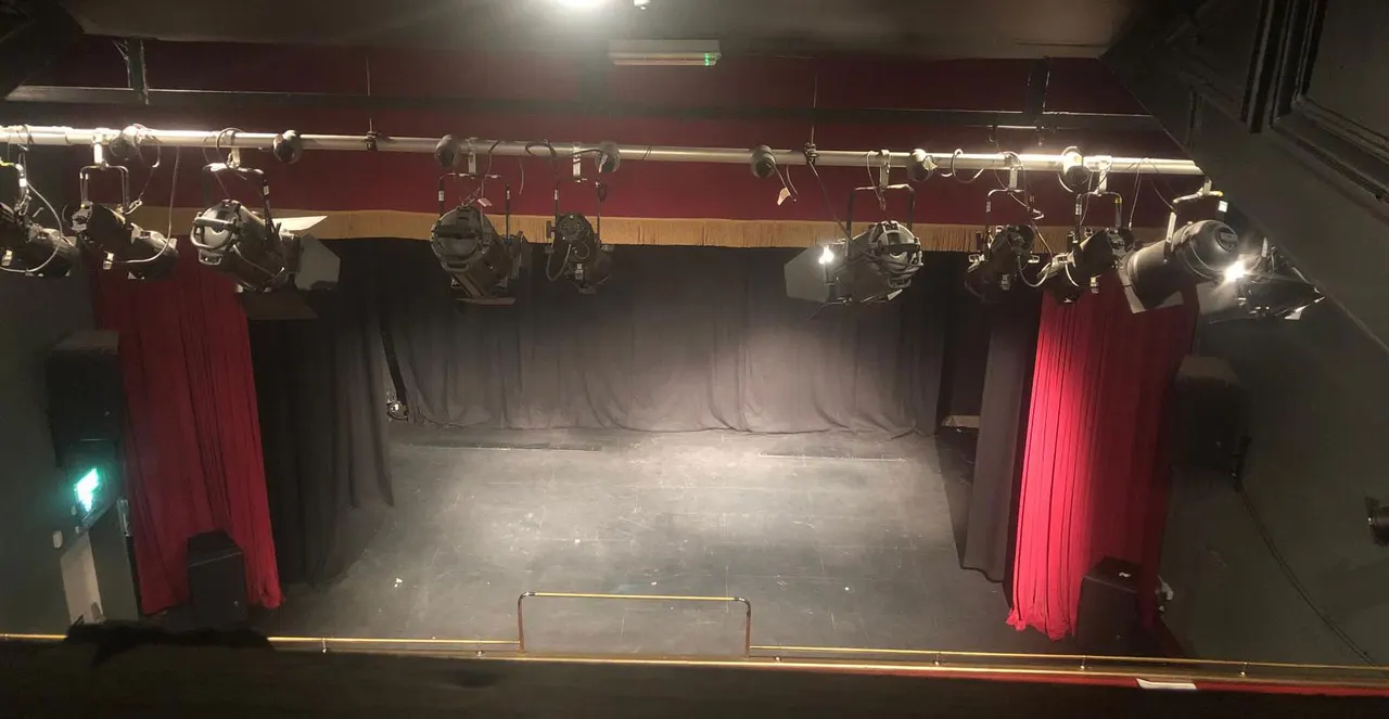 Empty theatre stage with red velvet curtains, professional lighting rigs overhead, and dark backdrop.