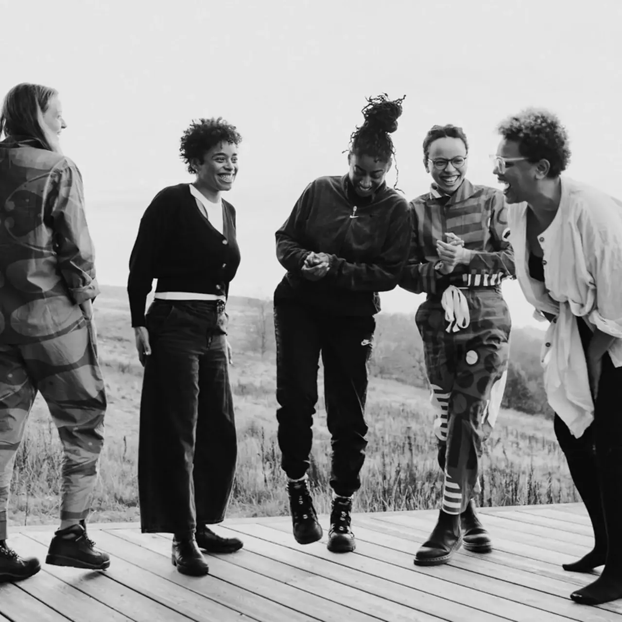 Five people gathered on a wooden deck overlooking rolling hills, laughing and chatting together in black and white.
