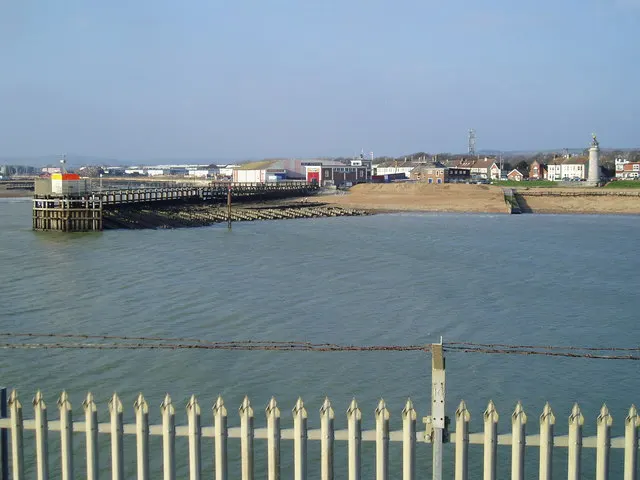 White picket fence in foreground with view across bay towards a pier, beach and seafront buildings under clear sky.