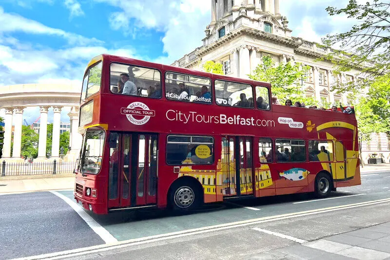 A red double-decker tour bus with passengers on a street in front of a classical stone building with columns.