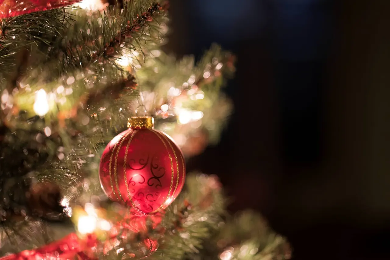 Red bauble with gold detailing hanging on a Christmas tree with twinkling lights.