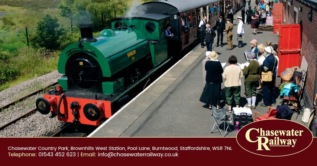 A green heritage steam locomotive at platform with visitors in period costume boarding carriages.