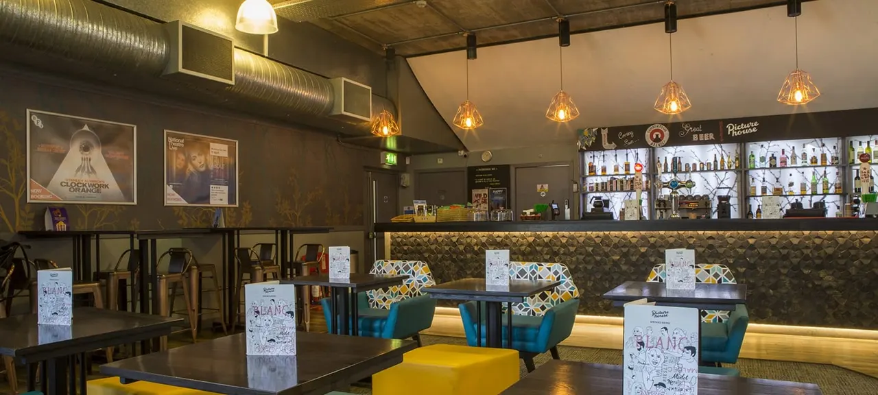 Modern bar interior with dining tables, teal chairs, hanging amber pendant lights, and illuminated spirit bottles behind the counter.