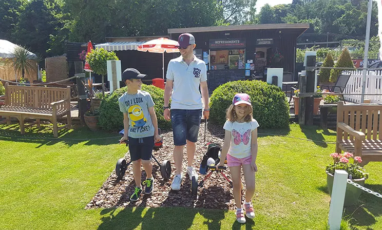 An adult and two children walk through a mini golf course with a food van and landscaped garden in the background.