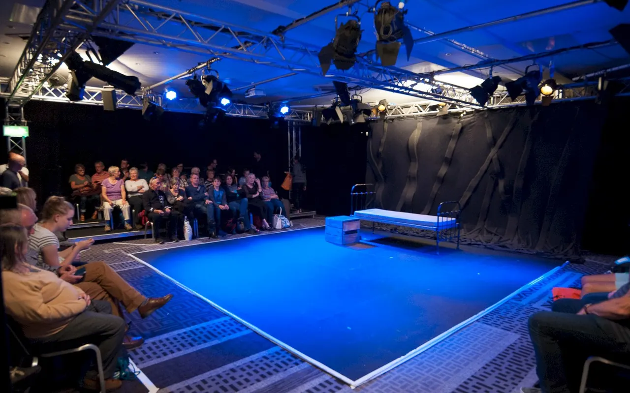 Audience members sit around a brightly lit blue circus or performance stage with professional rigging and lighting rigs overhead.