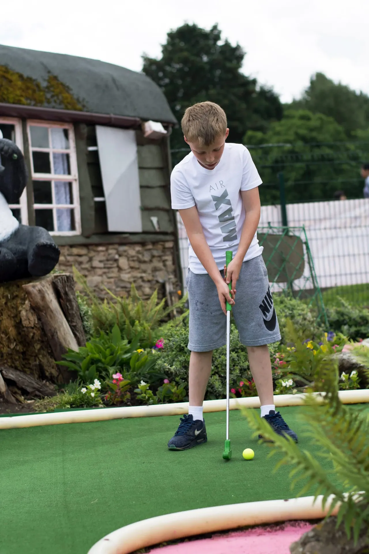 Child playing mini golf on a green course with a cottage and garden visible in the background.