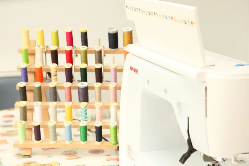 Wooden thread organiser with coloured spools of thread next to a sewing machine and design patterns.