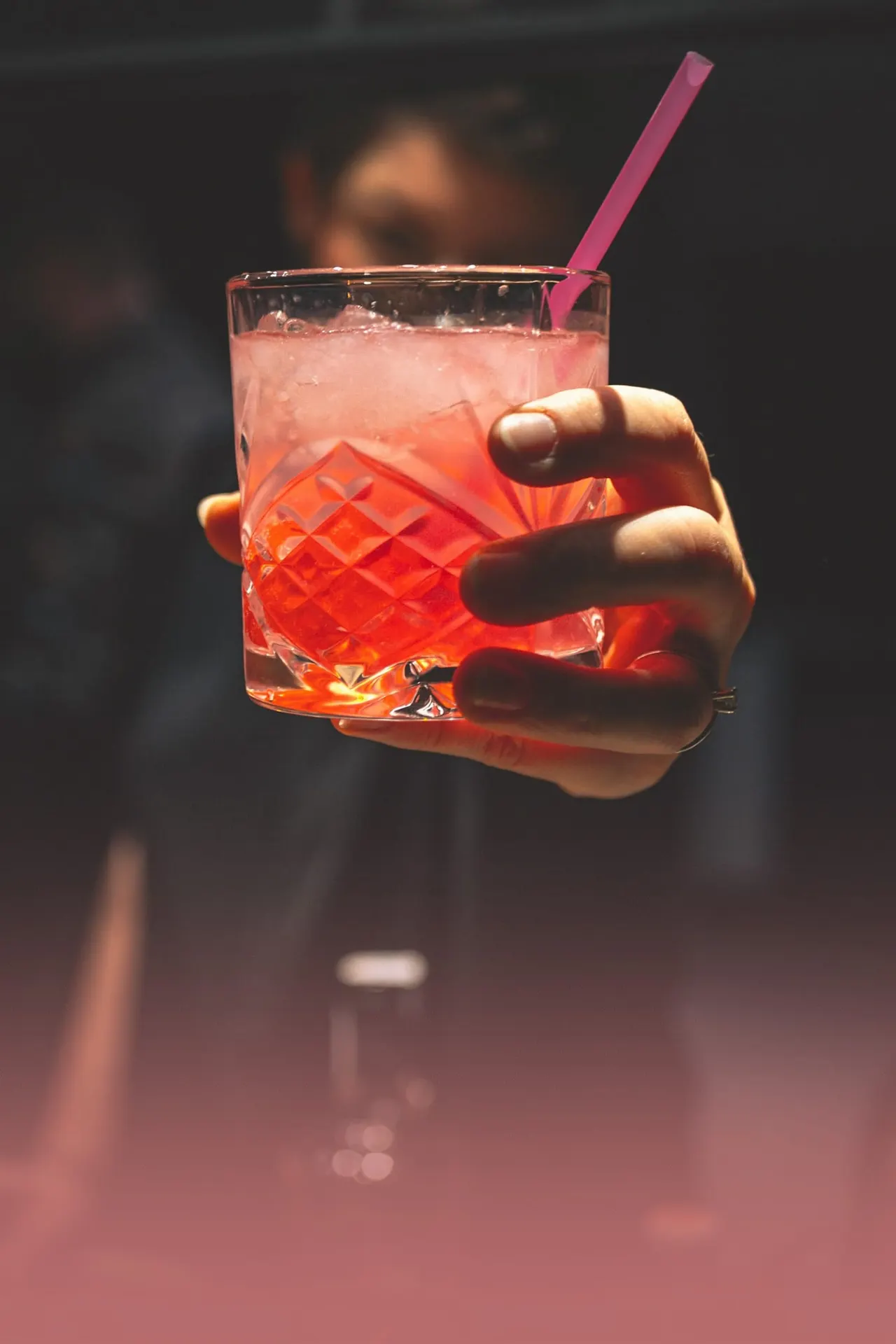 Hand holding a glass of red cocktail with ice and a pink straw against a dark blurred background.