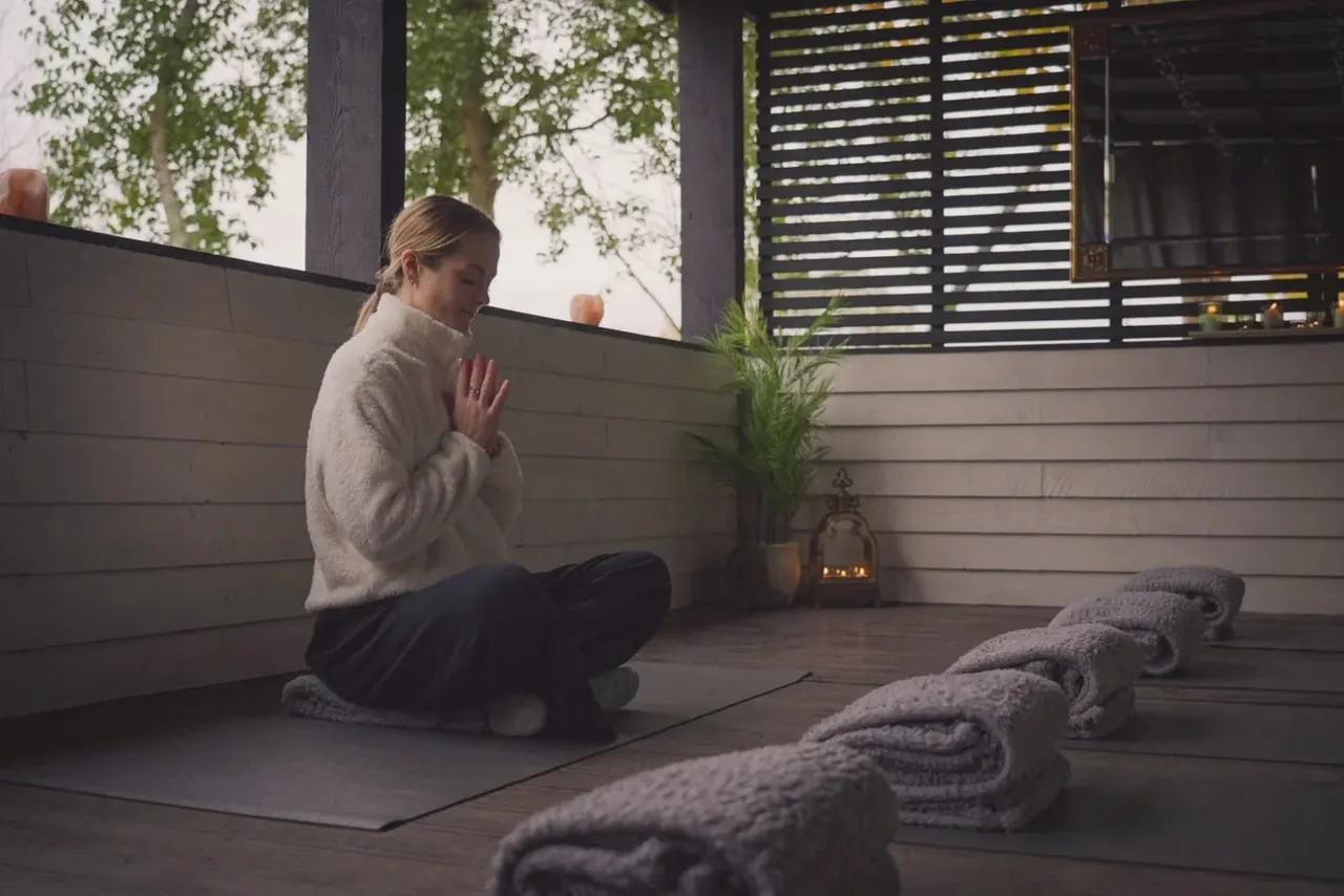 Woman in meditation pose on a yoga mat in a modern outdoor wellness pavilion with rolled towels nearby.