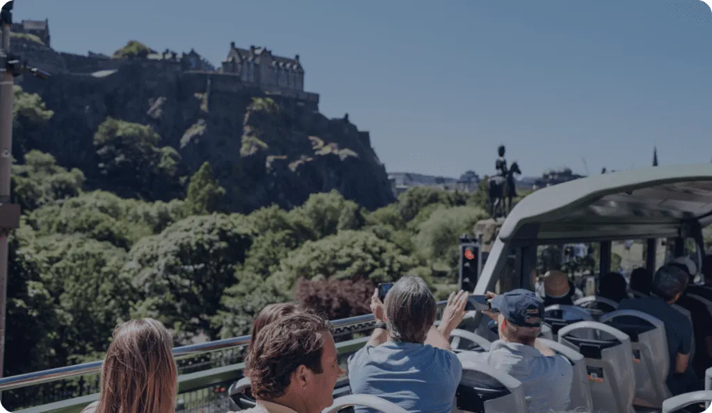 Tourists on an open-top bus with Edinburgh Castle perched on a rocky cliff in the background.