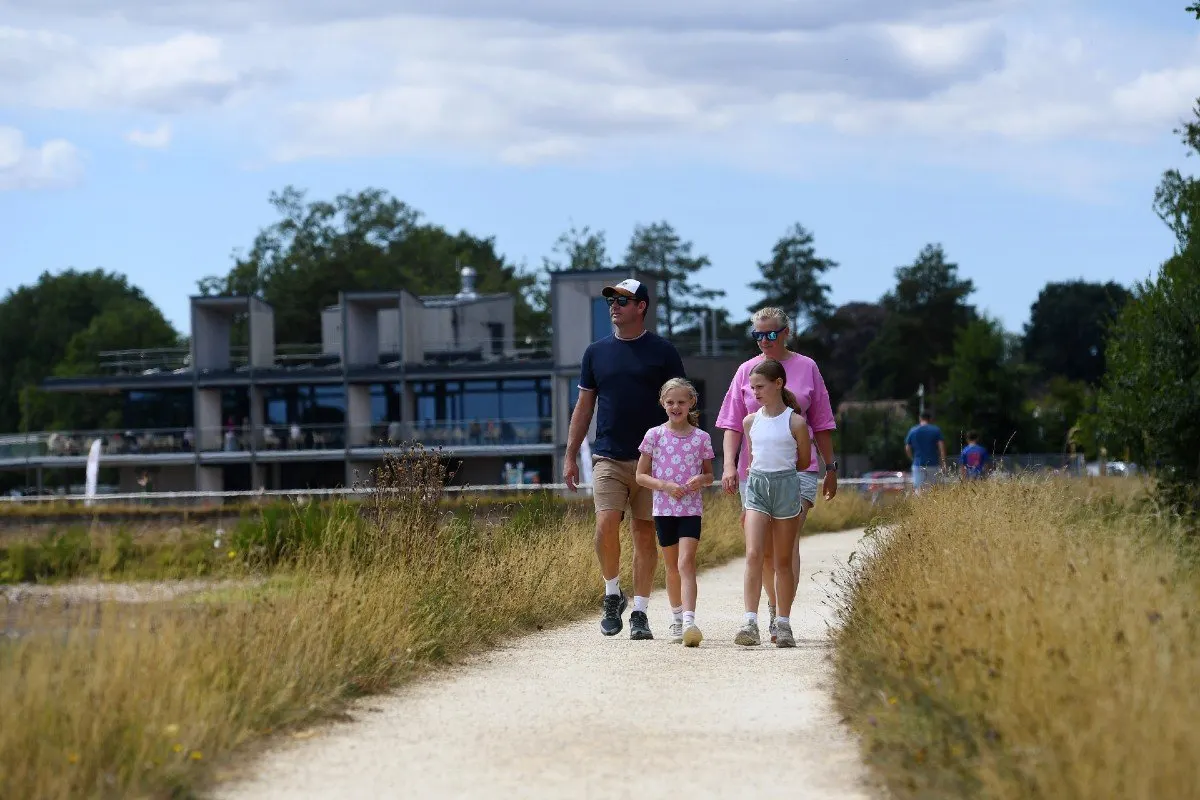 A family of four jogging along a sandy path beside dry grass, with a waterside building visible in the background.