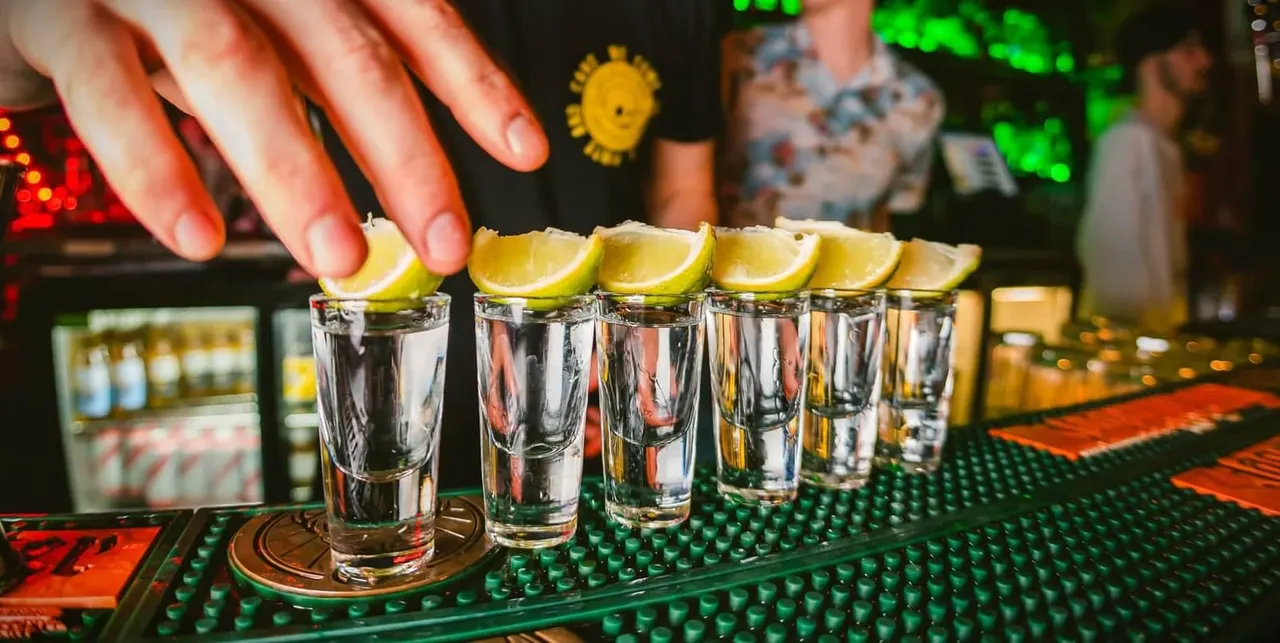 Hand placing lemon wedges on shot glasses filled with clear spirit at a busy bar counter.