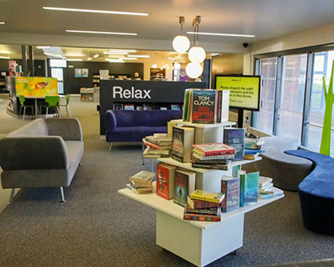 A modern library lounge area with comfortable seating, book displays, and a "Relax" sign above a blue sofa.