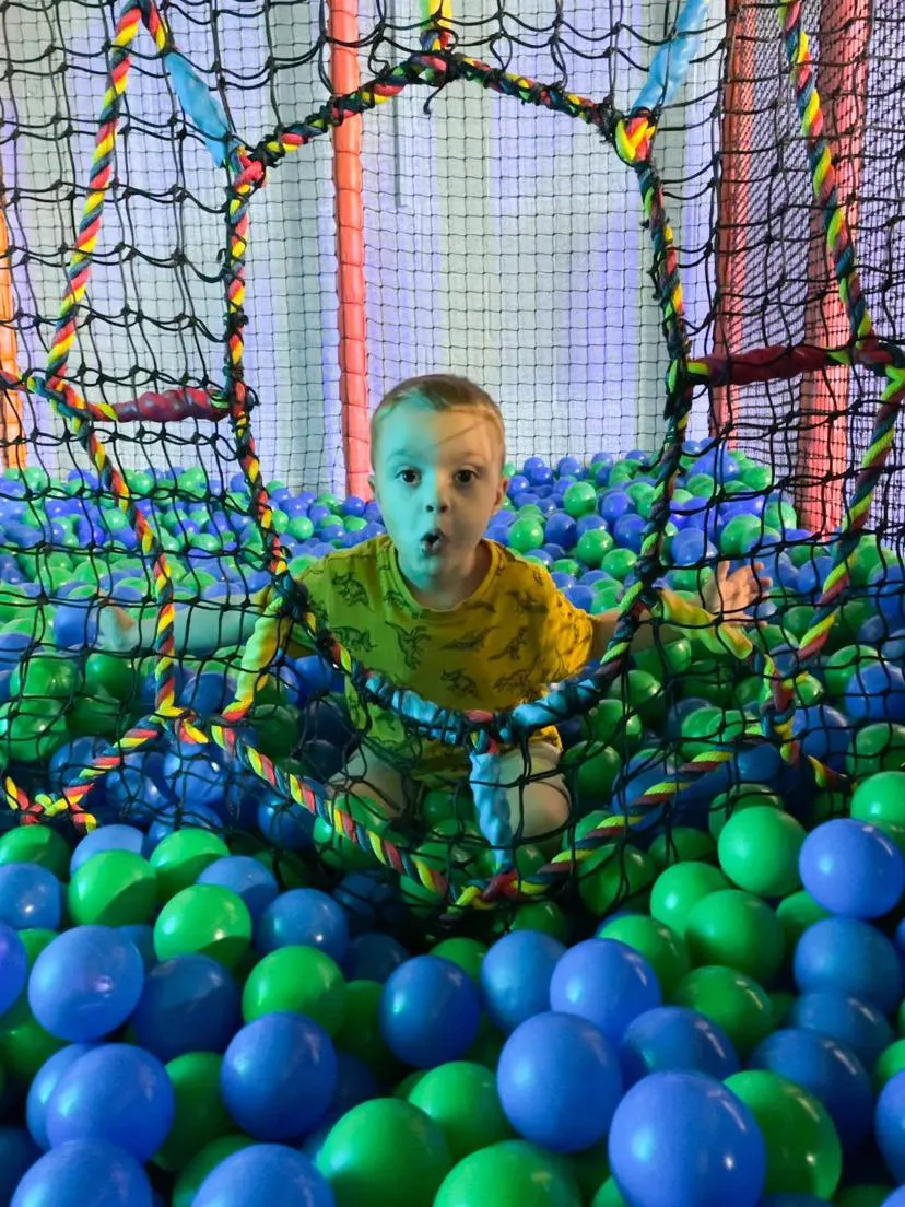 Young child in a yellow shirt standing in a colourful ball pit with blue and green balls, surrounded by netted play structures.