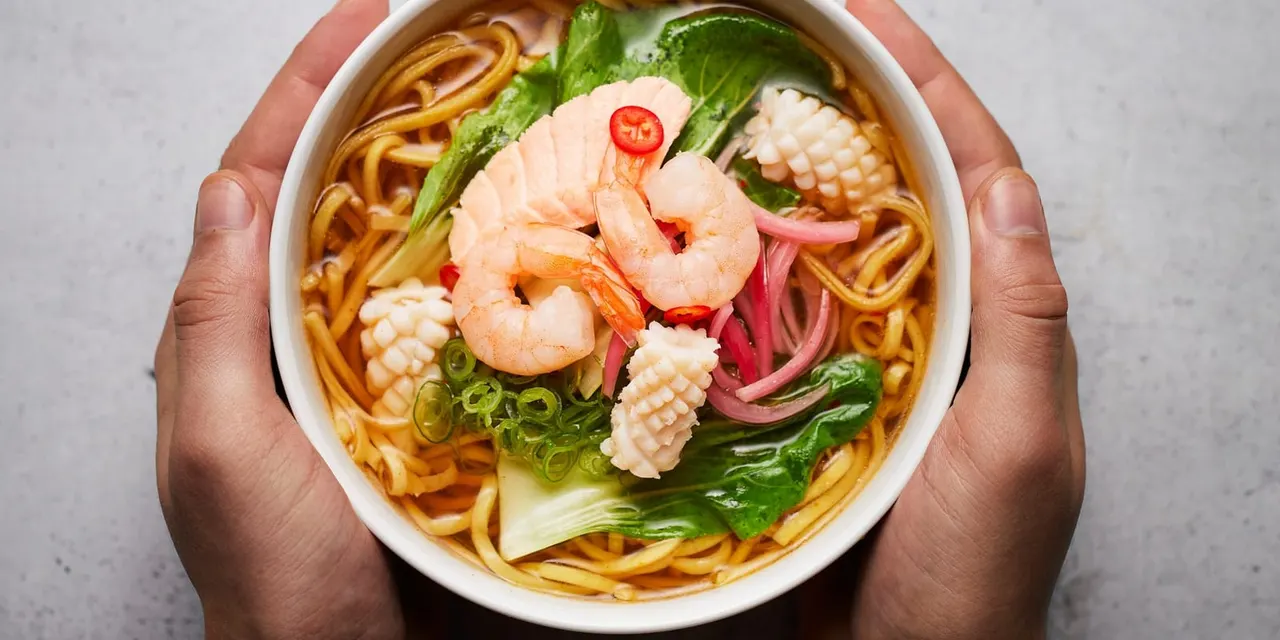 Hands holding a white bowl of noodle soup topped with prawns, octopus, greens and red chilli.