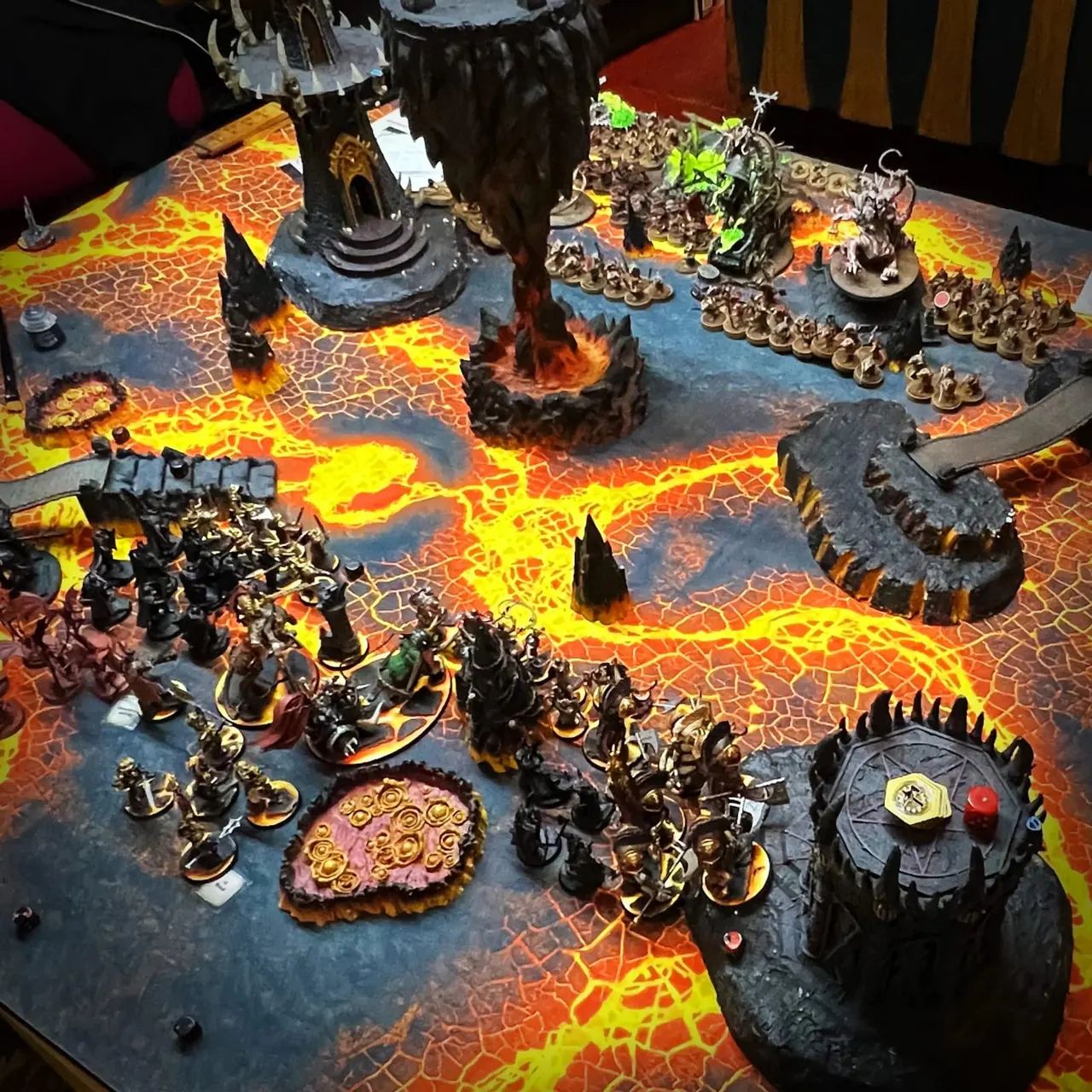 Tabletop wargaming set-up with painted miniature figures, terrain features and lava-effect board featuring dark gothic structures.
