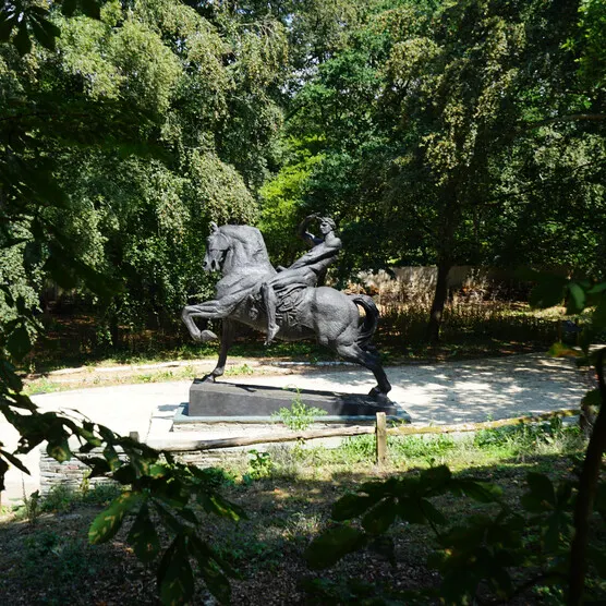 Statue of a rearing horse with rider in a wooded garden setting, surrounded by mature trees.