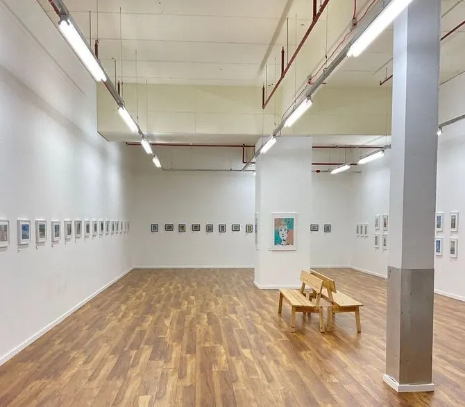 Gallery interior with wooden floor, white walls displaying small framed artworks, wooden benches, and overhead lighting.