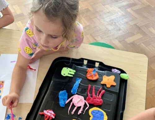 Young child leaning over a tray of colourful play dough models and figures at a craft activity table.