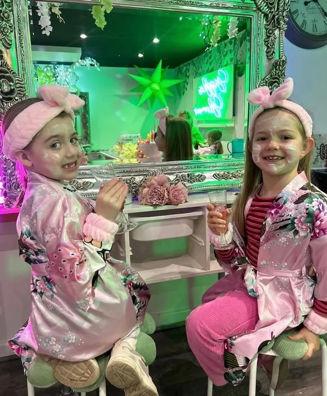 Two young children in pink spa robes and headbands sit at a brightly lit vanity with neon green accents, enjoying a glamour activity together.