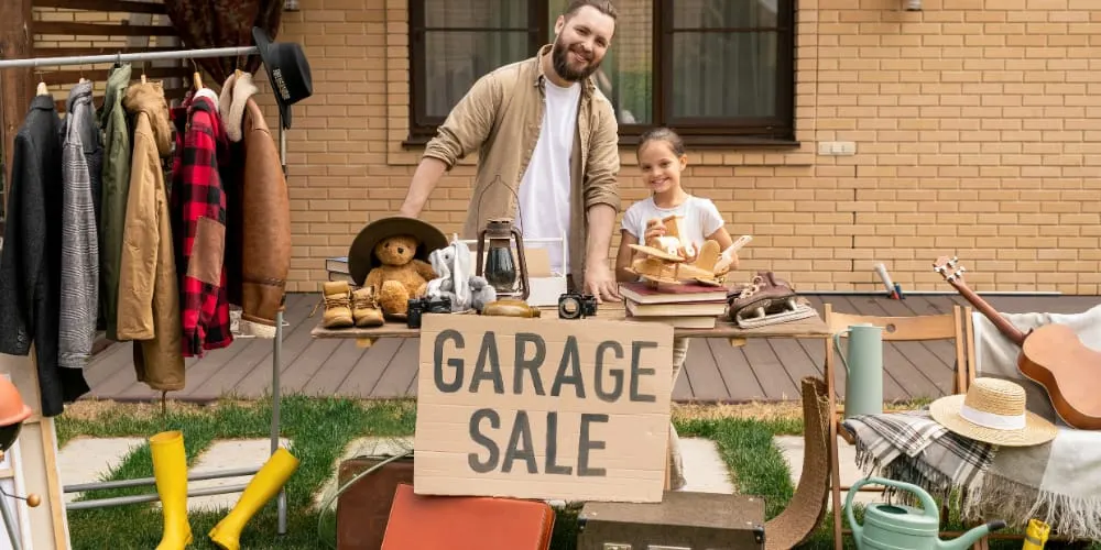 Adult and child at a garage sale table displaying second-hand clothing, toys and household items outside a brick building.