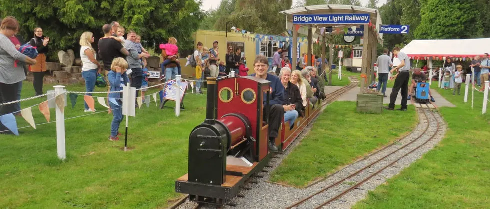 Visitors riding a miniature train at an outdoor railway attraction, with families watching from the grassy surroundings.