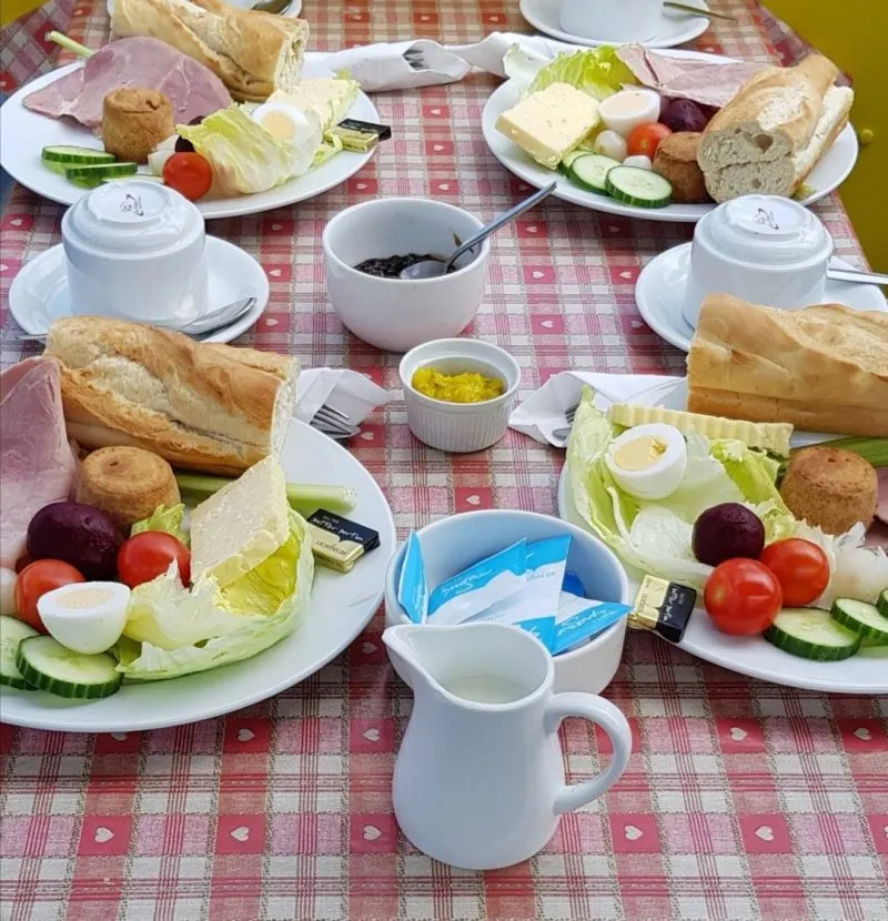 Overhead view of a ploughman's lunch spread on a red and white checkered tablecloth with bread, cheese, cured meat, fresh vegetables and beverages.