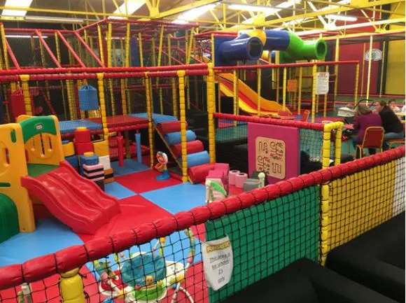 Children play in a colourful indoor soft play area with slides, climbing structures, foam blocks and padded equipment.