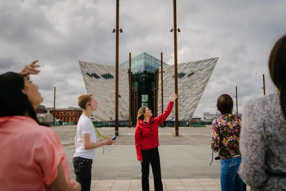 A tour guide in a red jacket points towards the Titanic Belfast building whilst leading a group of visitors across the outdoor plaza.