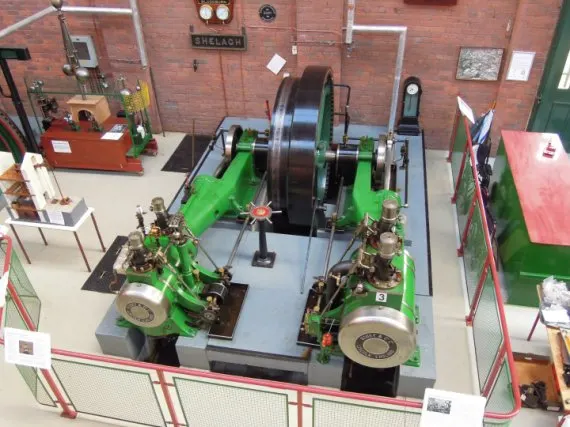 Overhead view of a large green steam engine on display with two smaller engines flanking it, inside a brick museum building.
